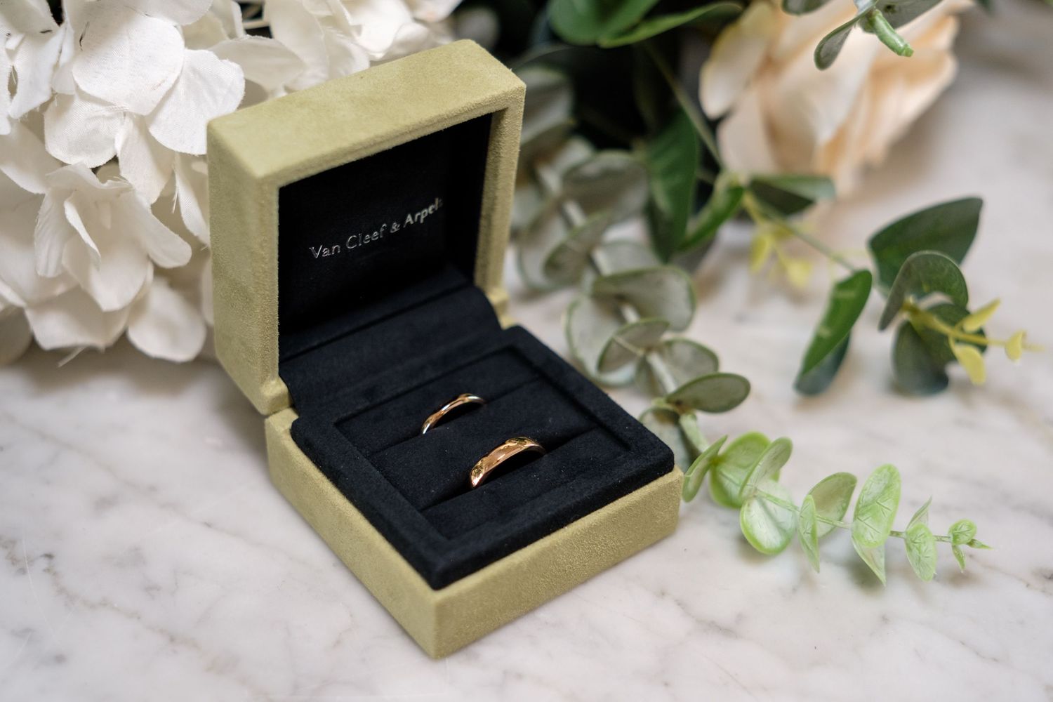 Wedding rings nestled in black velvet boxes surrounded by delicate white flowers and eucalyptus on a marble surface.