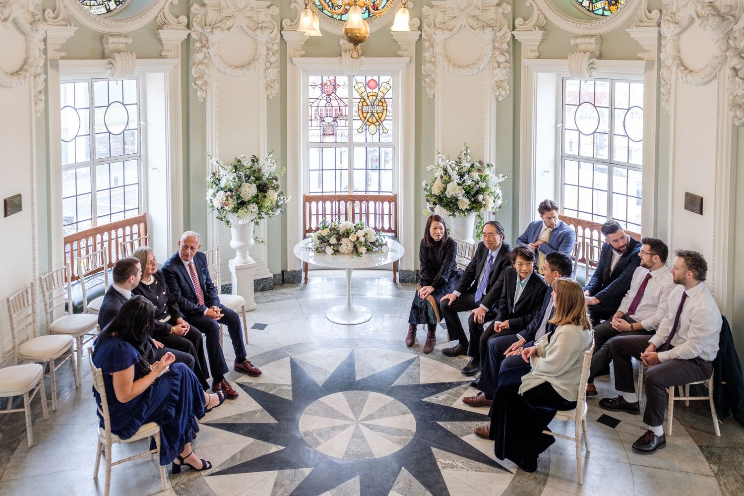 Wedding guests gather in an elegant white room with ornate details and large windows for a celebration event.