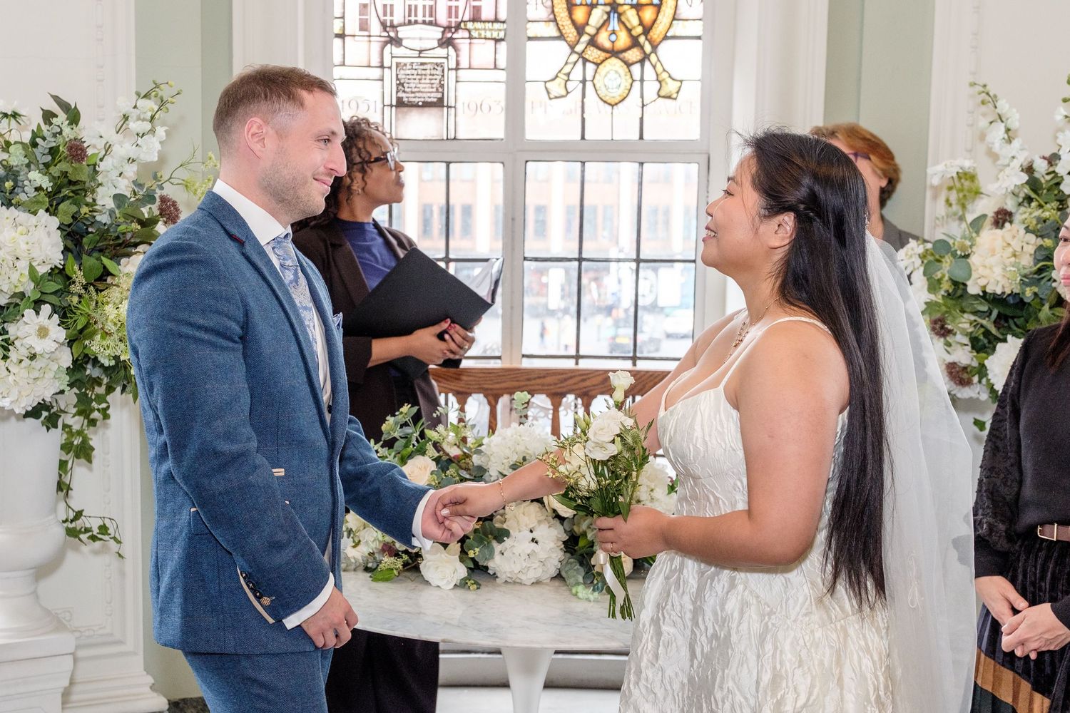 A couple exchanges vows during an indoor wedding ceremony surrounded by white floral arrangements and elegant decor.