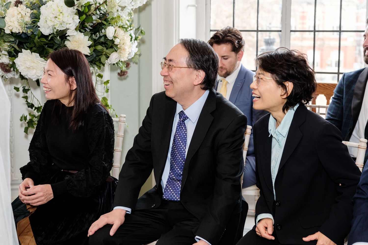 Wedding guests seated in rows share laughter during a joyful celebration at an indoor venue.