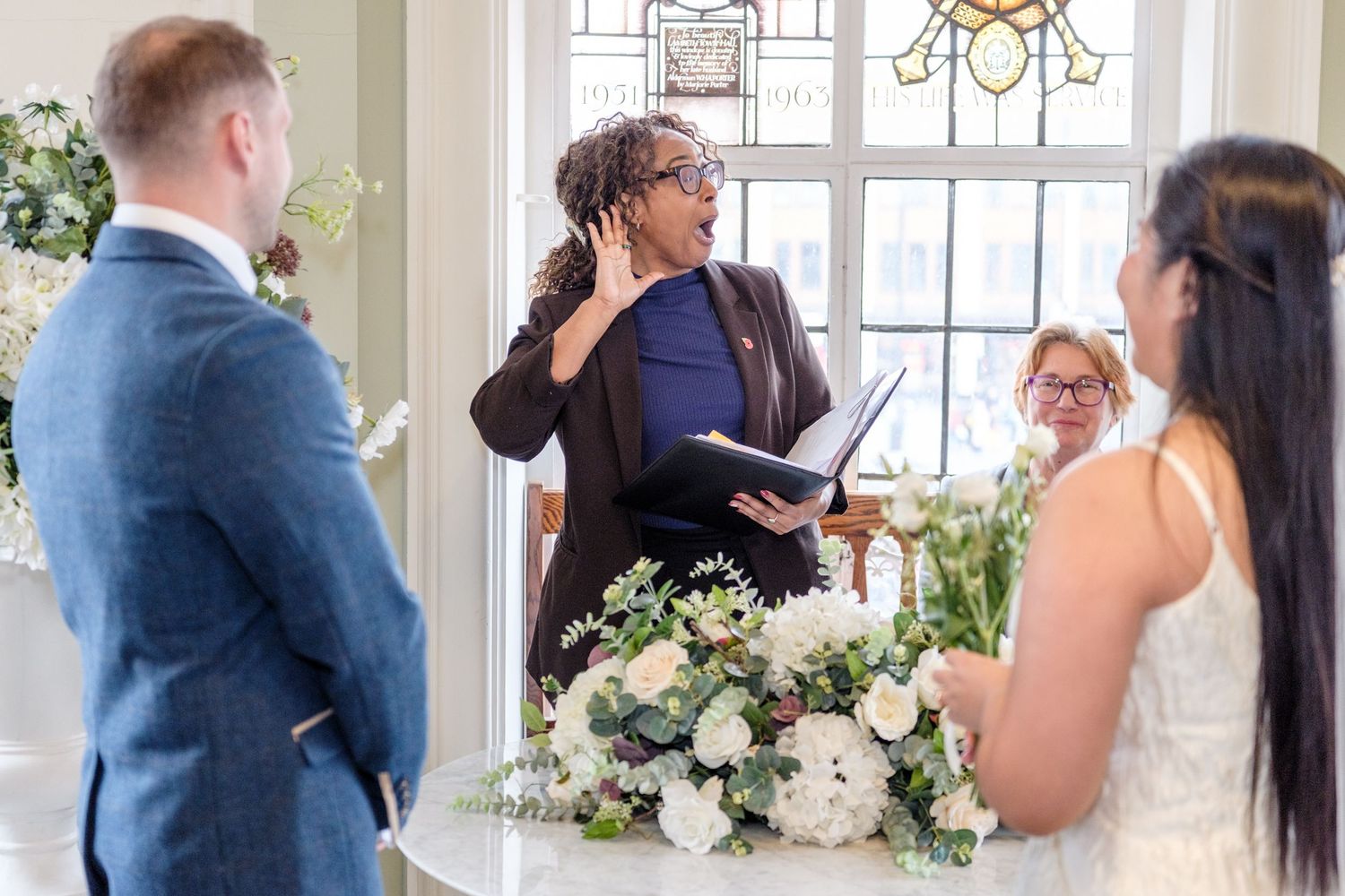 A wedding ceremony takes place indoors with an officiant and couple standing near elegant white floral arrangements.