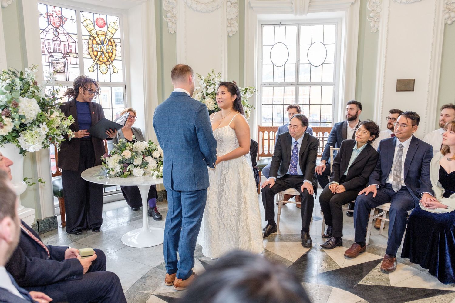 A wedding ceremony takes place in an elegant white room with ornate windows and floral arrangements.