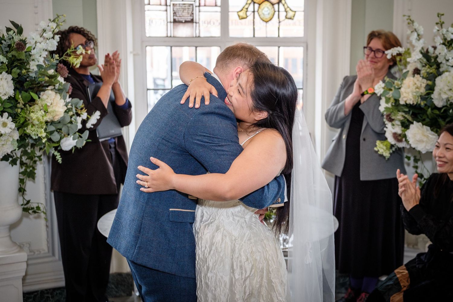 A tender embrace during a wedding ceremony with guests looking on in an elegant white room.