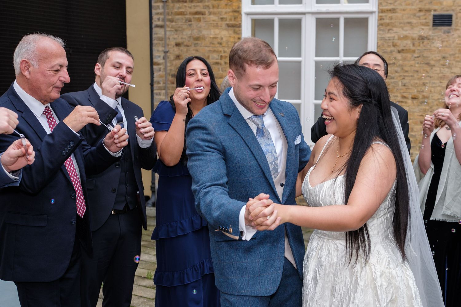 Wedding party laughs together during candid outdoor photo session at brick venue.
