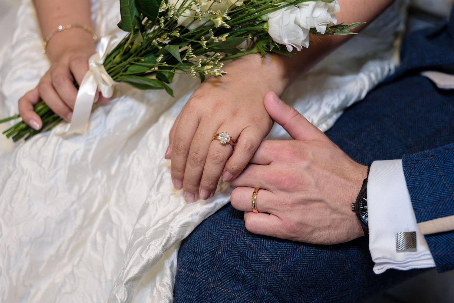 Close-up of hands exchanging wedding rings with white flowers and formal attire visible in the frame.