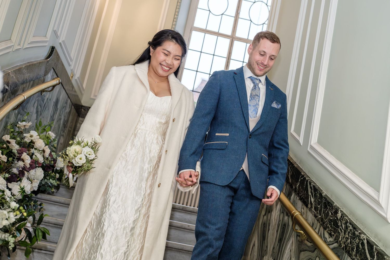 Wedding couple poses in elegant indoor venue with white floral arrangements and classic architectural details.