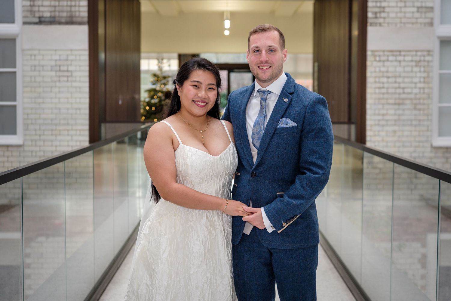 A couple in wedding attire poses together in a modern hallway with white brick walls and glass panels.