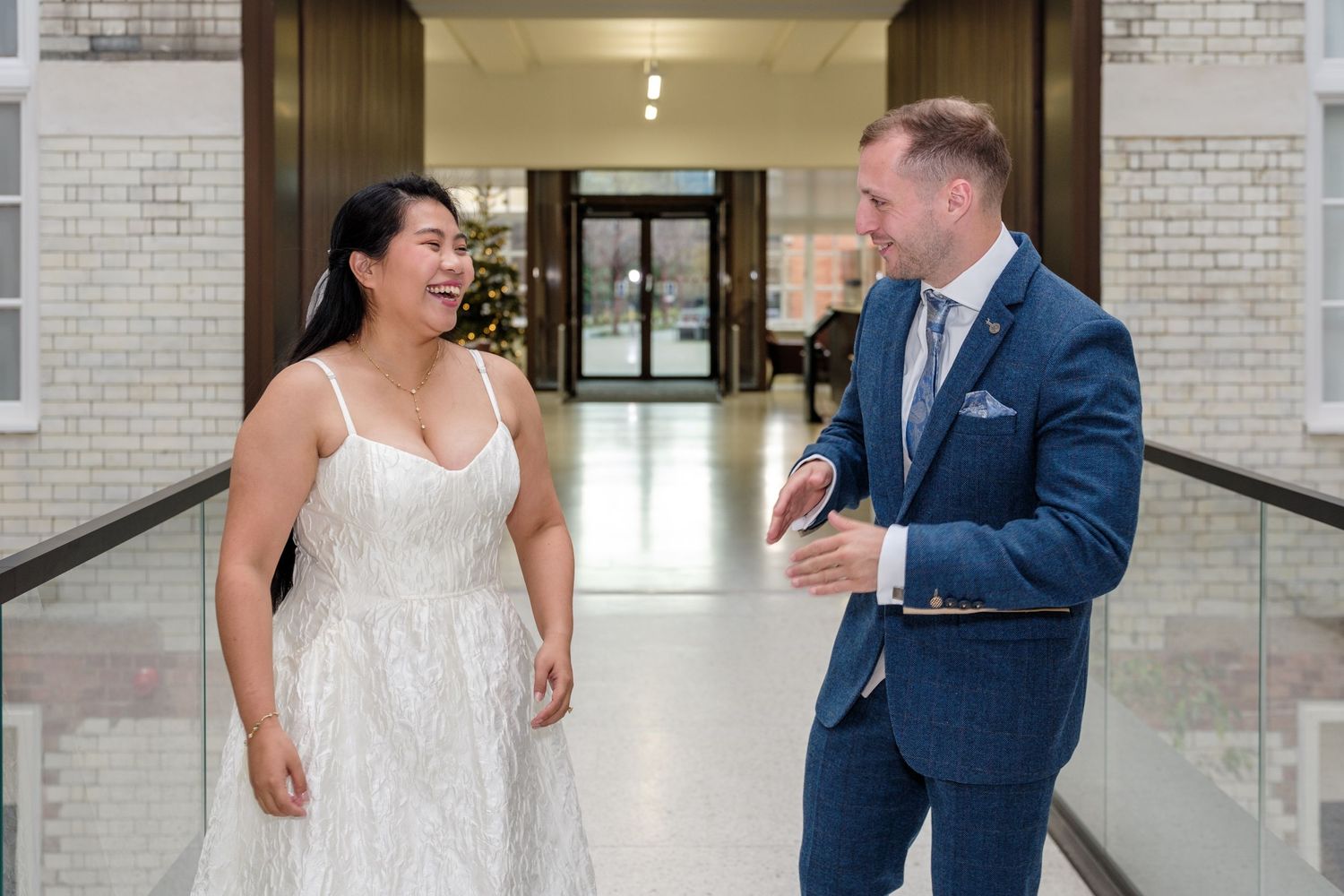 A couple in wedding attire laughs together in a bright indoor venue with brick walls and glass railings.