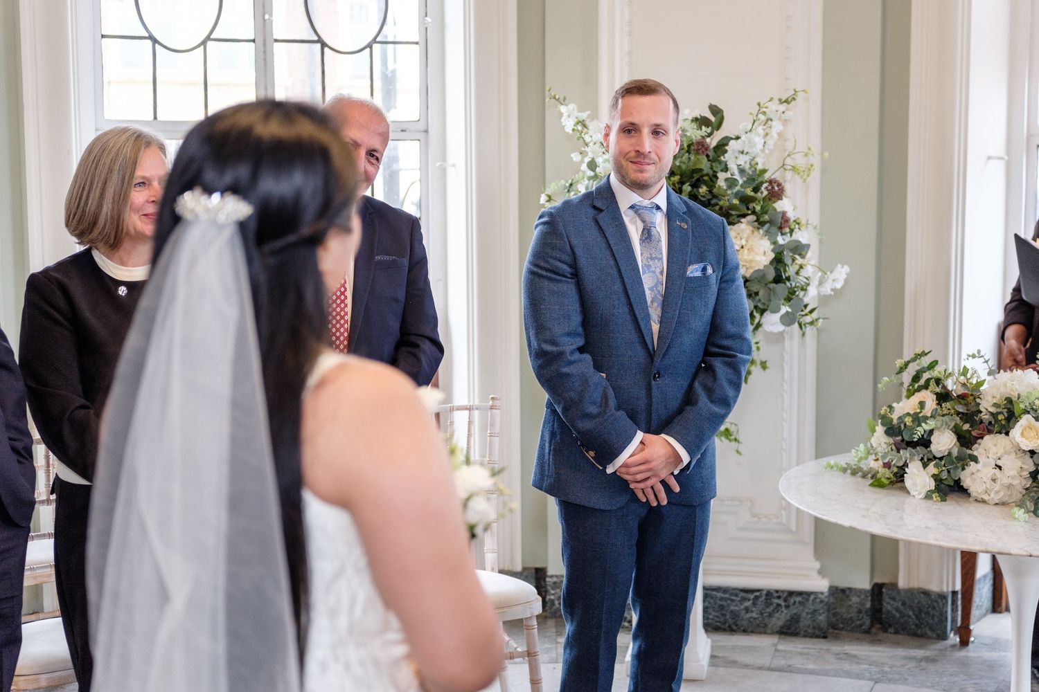 Wedding ceremony taking place in an elegant white room with guests standing and watching the couple exchange vows.