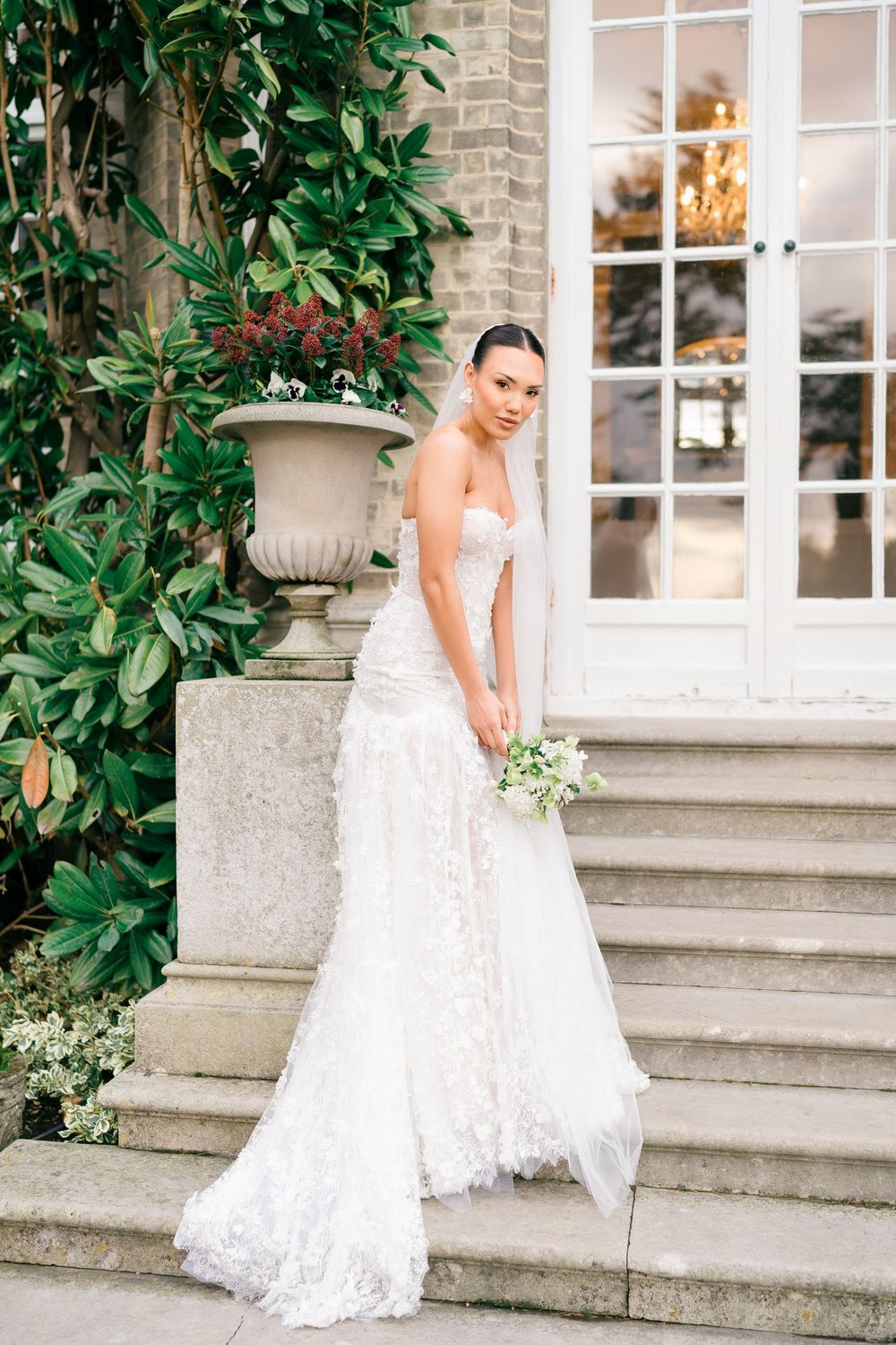 A bride in a white lace wedding dress poses gracefully on stone steps next to lush green foliage and white windows.