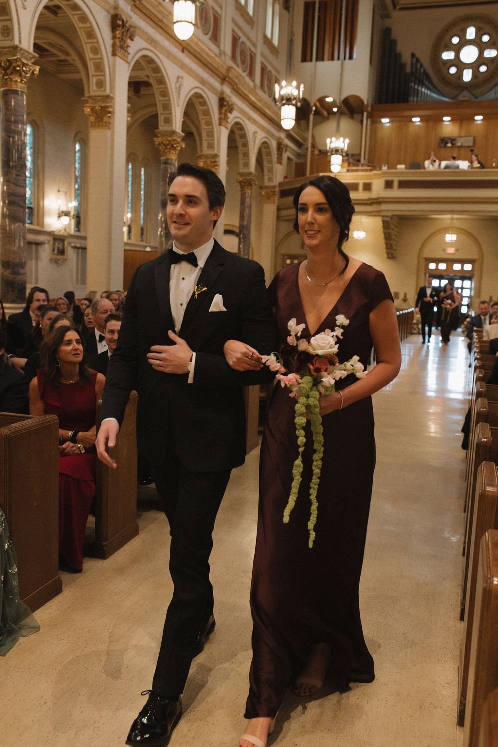 Wedding party members in dark formal attire walk down an elegant church aisle with ornate architectural details.