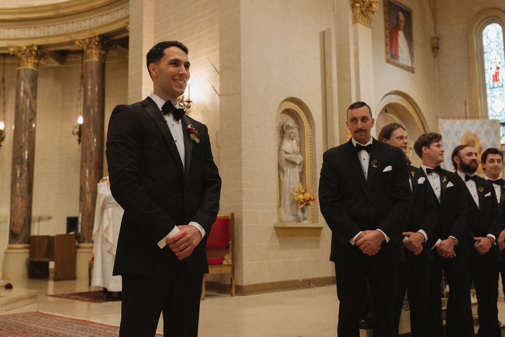 Groomsmen in black tuxedos stand in line at the altar of an ornate church during a wedding ceremony.