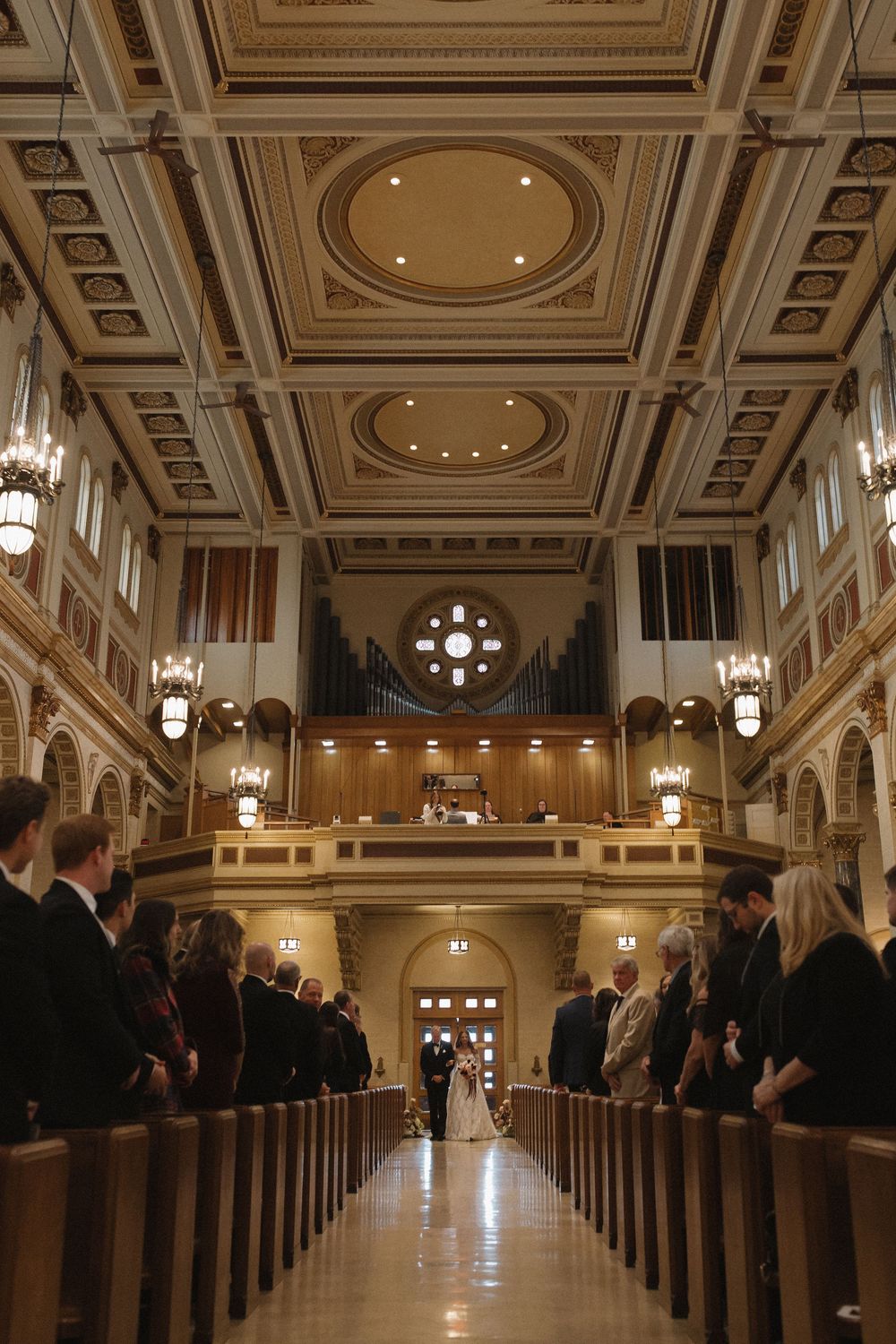 An elegant church interior with rows of wooden pews and hanging chandeliers during a wedding ceremony.
