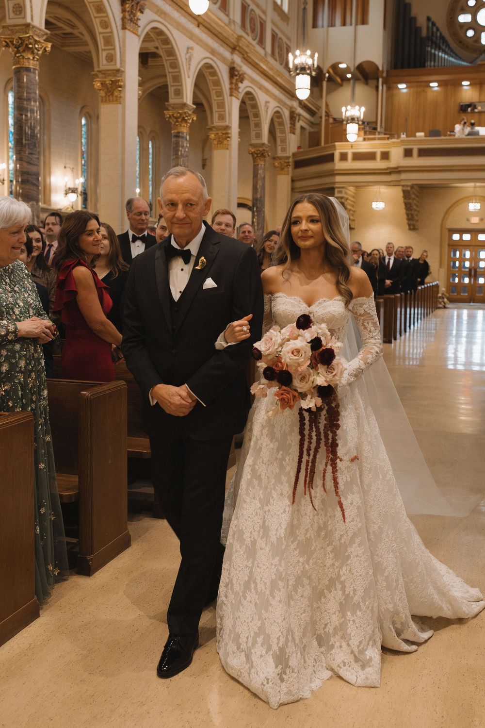Wedding party walks down the ornate church aisle with beautiful white floral arrangements and golden decor.