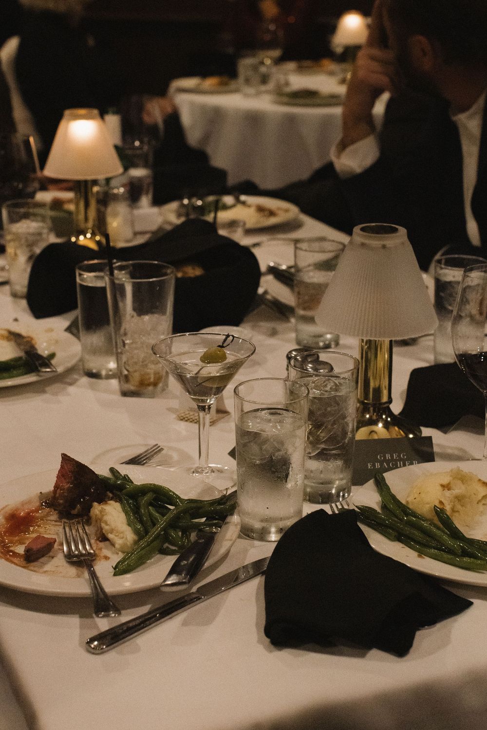 Elegant dining table settings at an upscale restaurant with white tablecloths, glassware, and green vegetables on plates.
