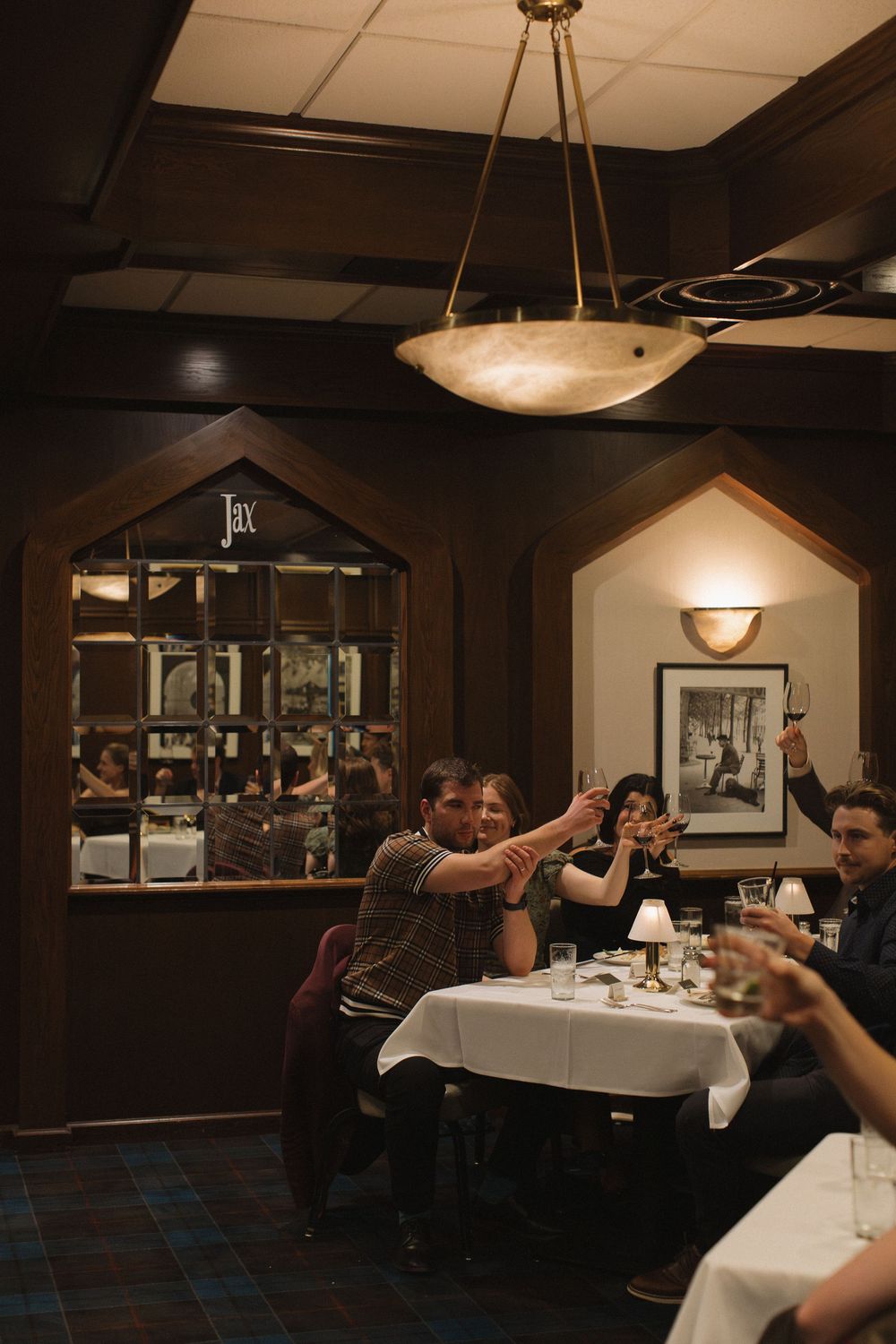 Diners seated at white-clothed tables in a dimly lit restaurant with pendant lighting and dark wood paneling.