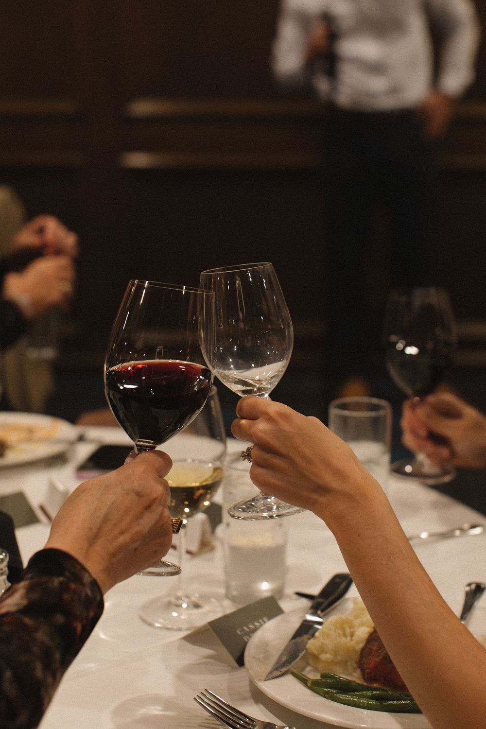 Diners holding wine glasses at an elegant restaurant dinner table with formal place settings.