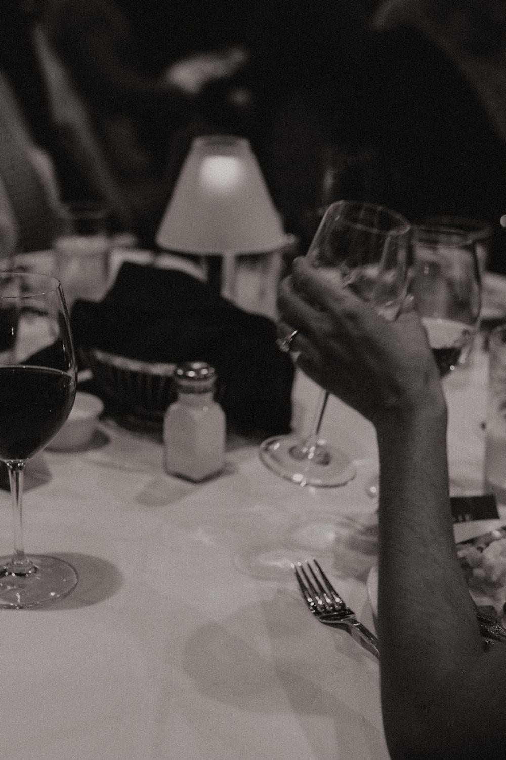 Black and white photo of hands reaching for wine glasses on an elegant dining table with white tablecloth.