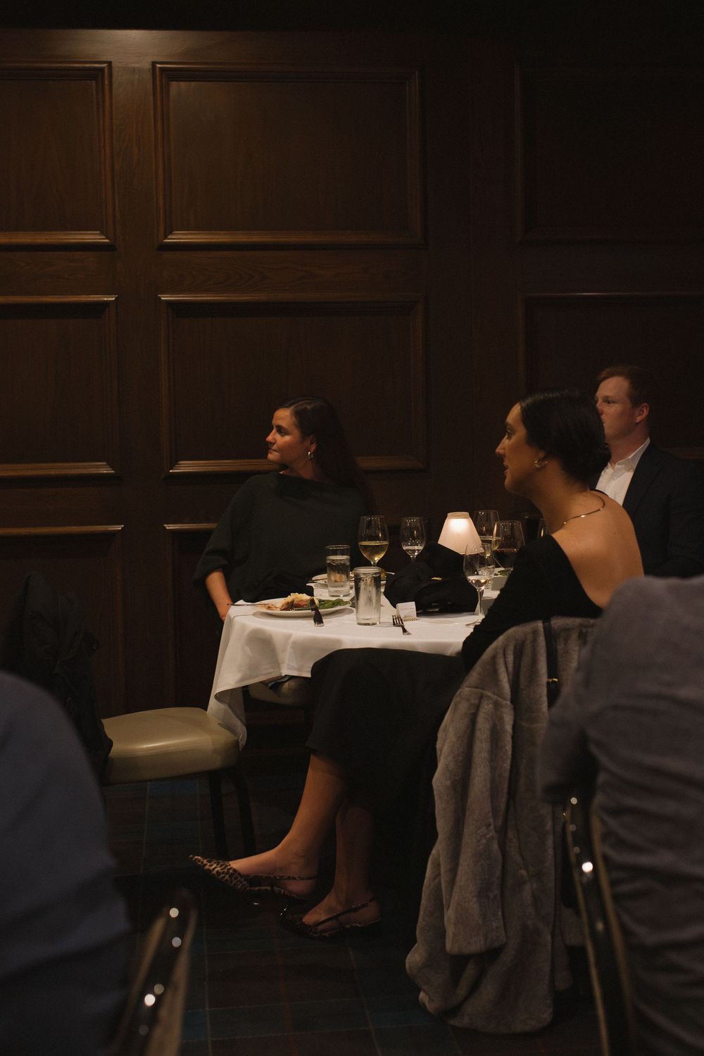 Diners seated at elegant tables in a dark wood-paneled restaurant with white tablecloths and wine glasses.