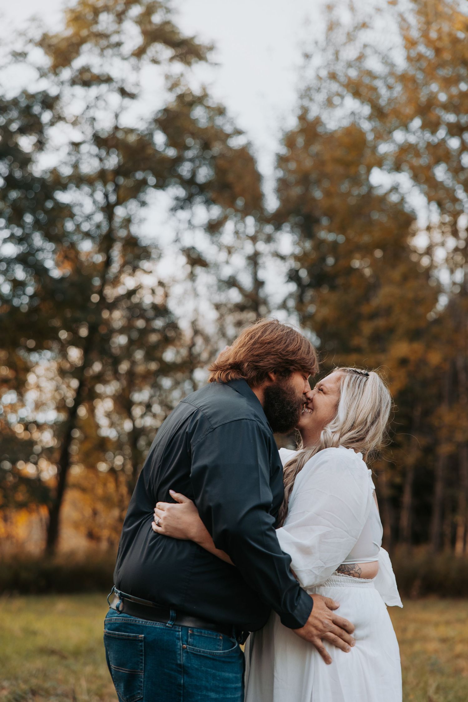 A romantic couple embraces and kisses in a fall forest setting with golden autumn trees in the background.
