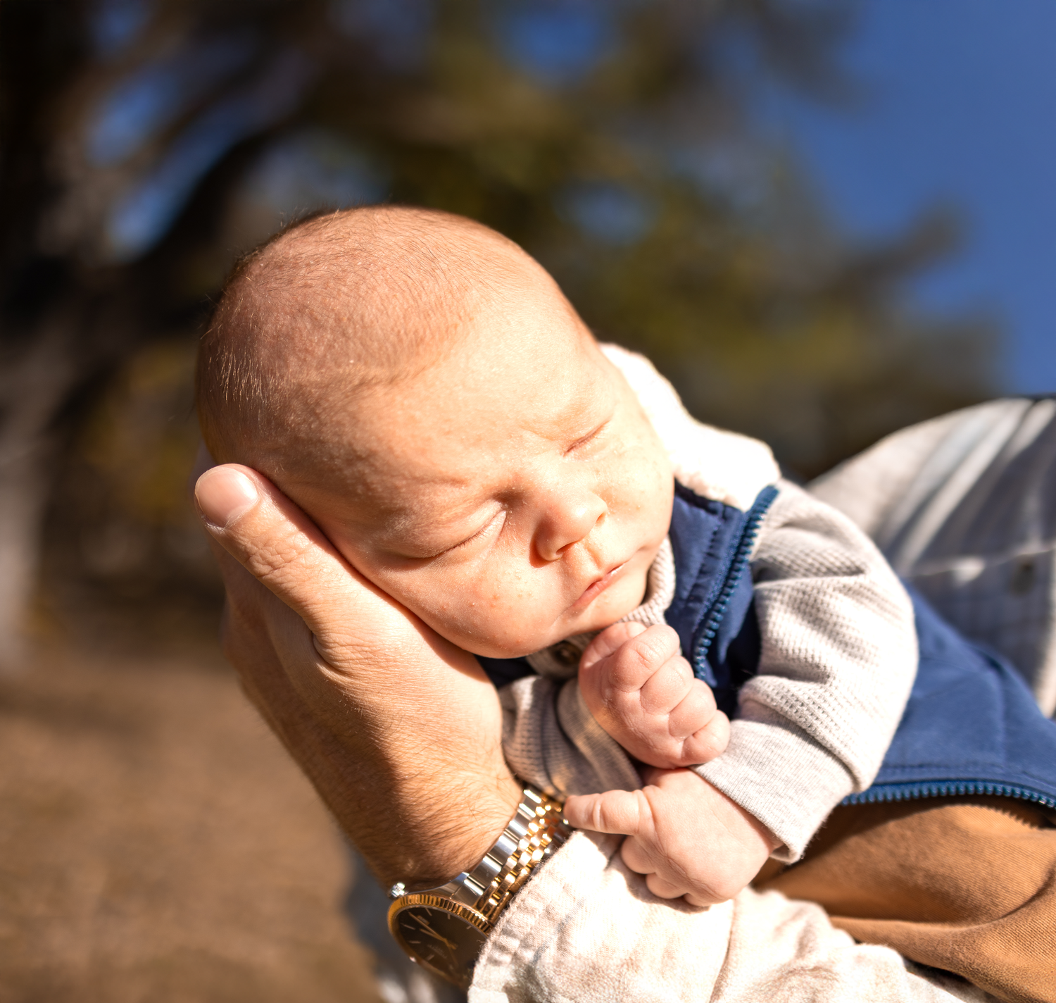 A sleeping newborn baby cradled gently in adult hands wearing a blue and gray outfit.