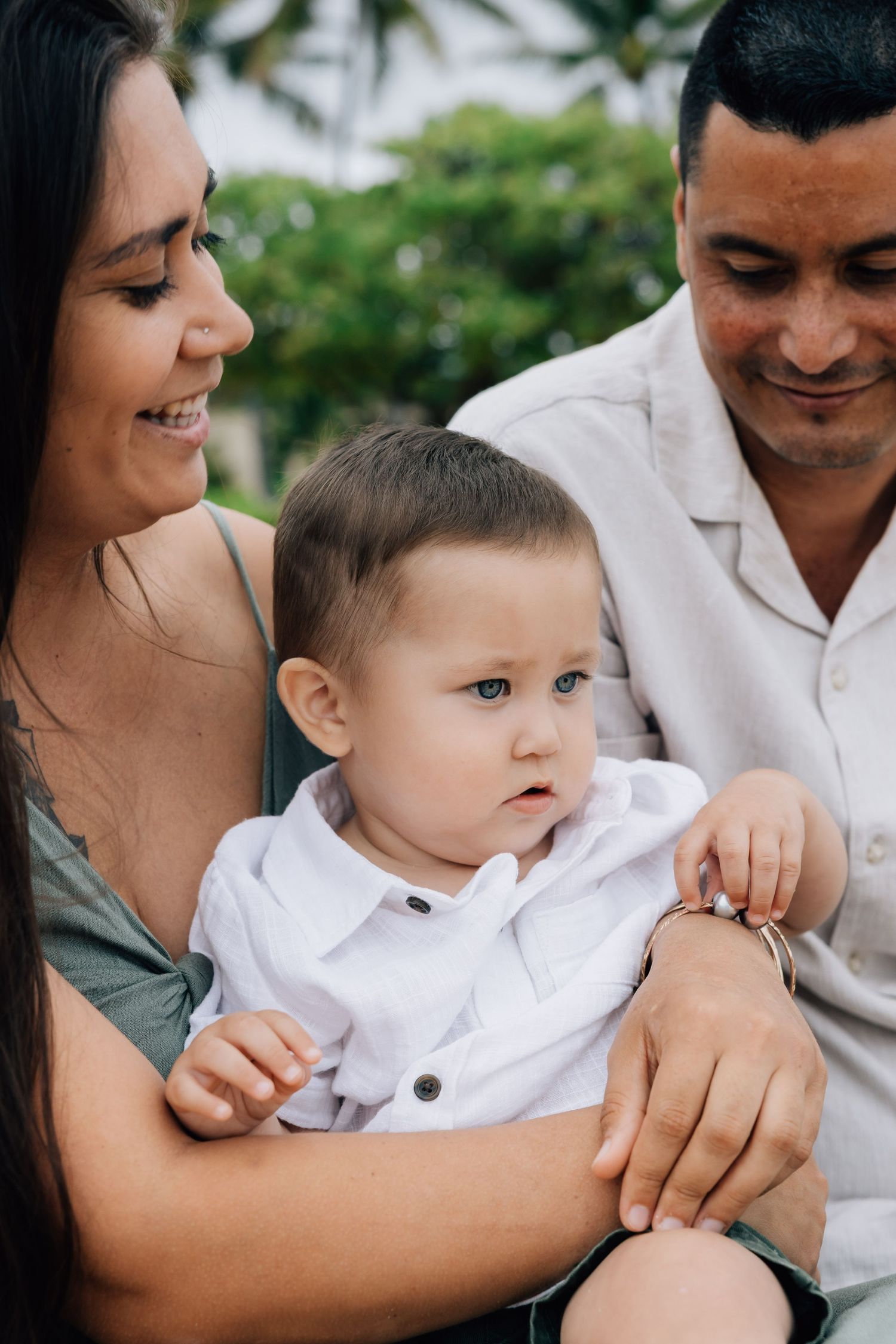 Loving parents smiling at their blue-eyed baby boy in a tropical setting.