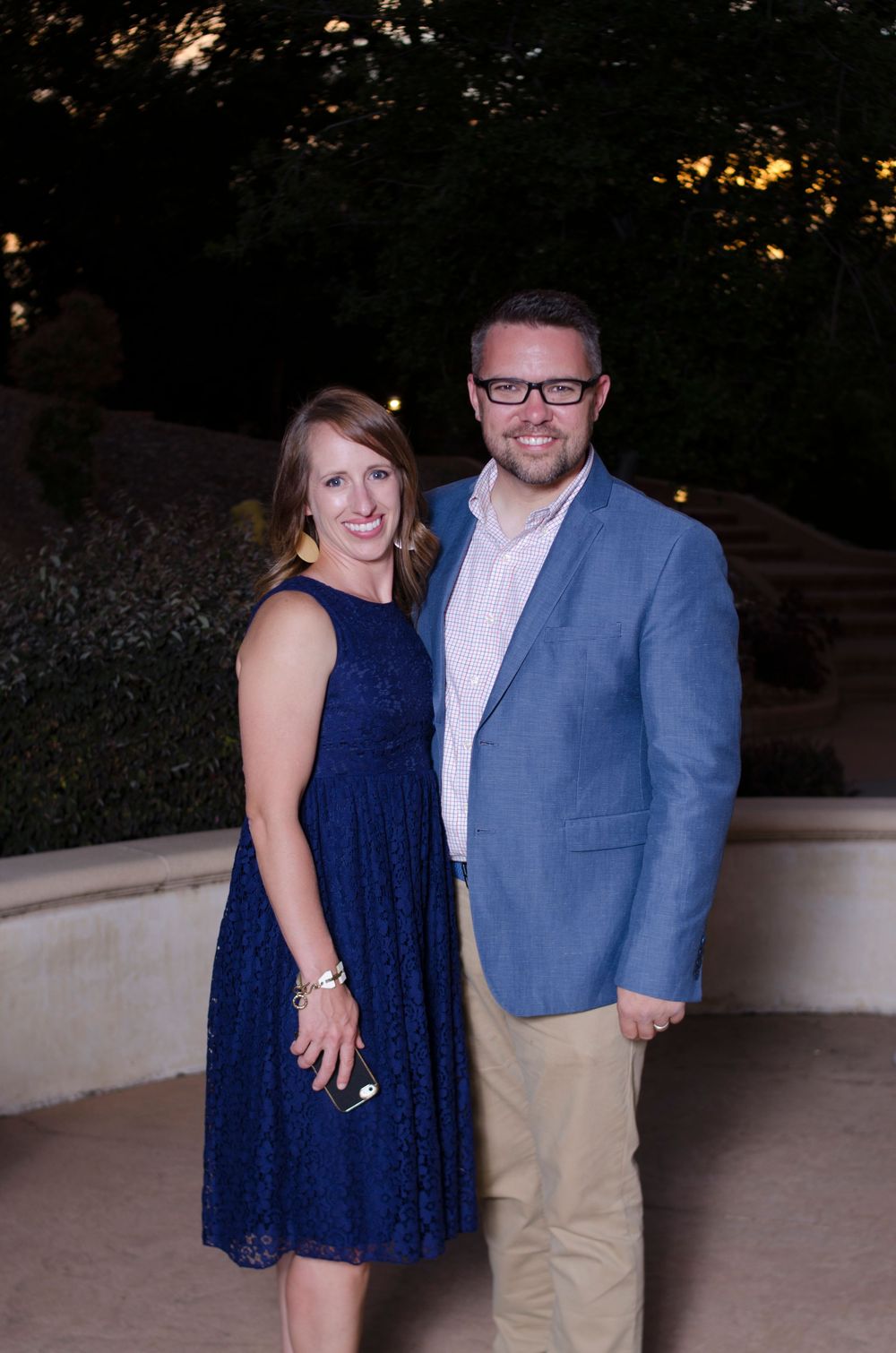 A couple poses together at an outdoor event, with the woman wearing a navy blue dress and the man in a light blue blazer.