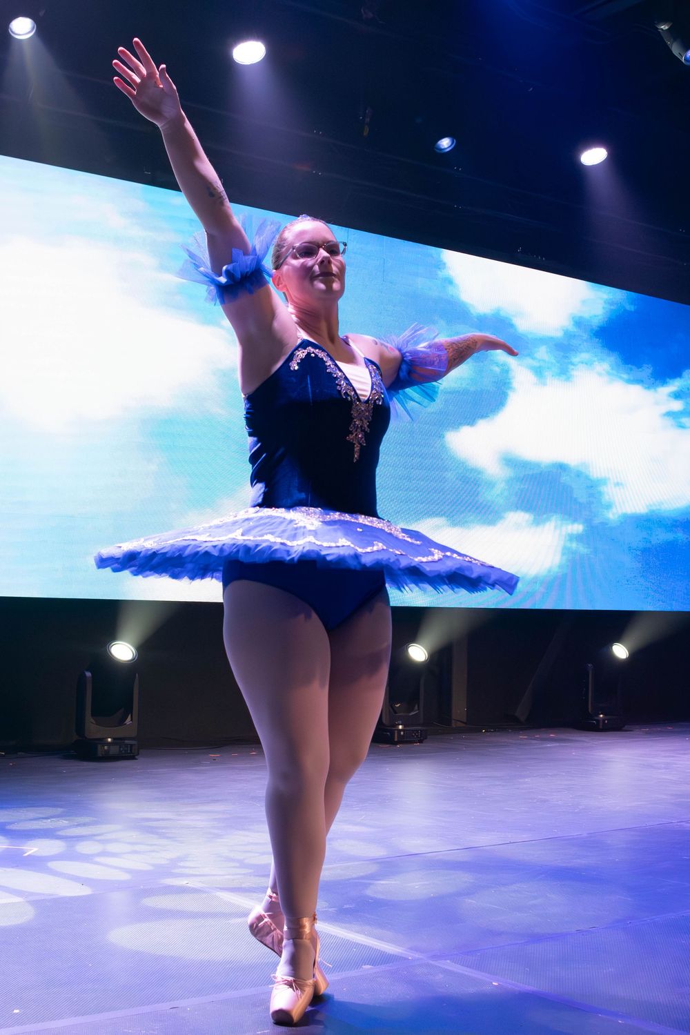 A dancer in a black leotard performs graceful movements on stage with blue lighting and sky backdrop.