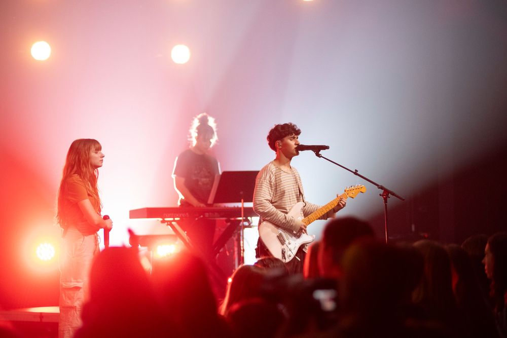 Rock band performs on stage with red lighting and fog effects creating a dramatic concert atmosphere.