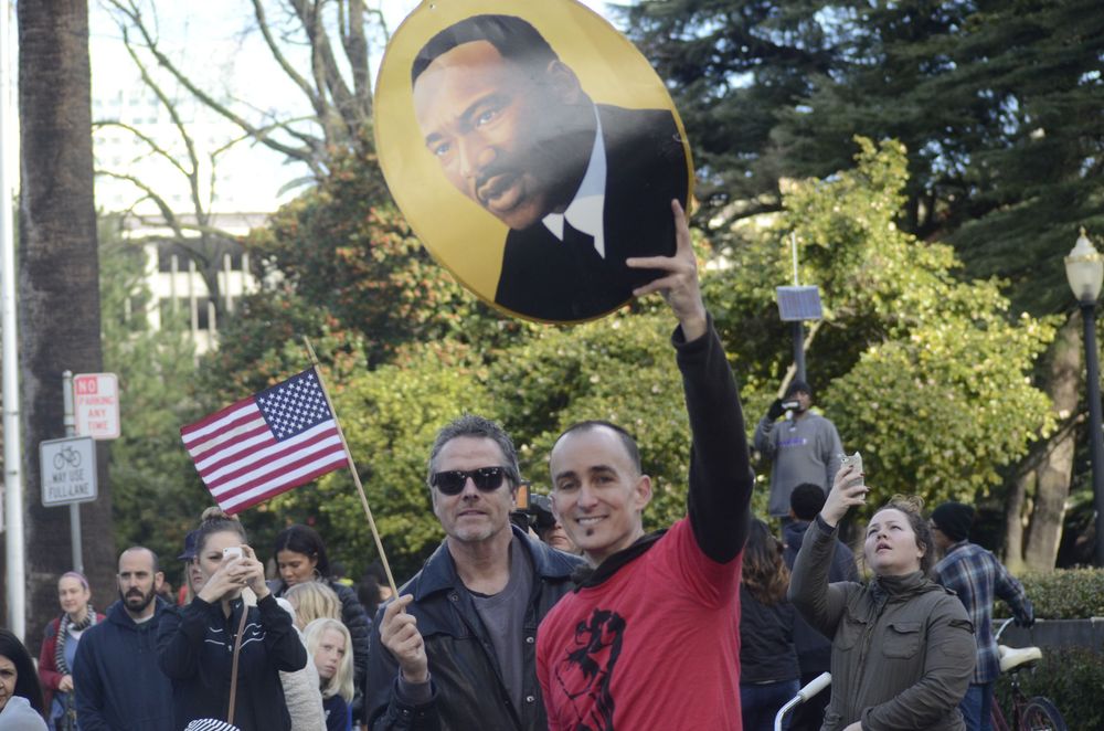 Protesters gather outdoors holding signs and an American flag during a public demonstration.