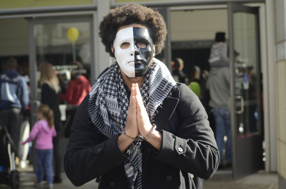Street performer wearing white face paint and black clothing performs with a handheld prop in an urban setting.