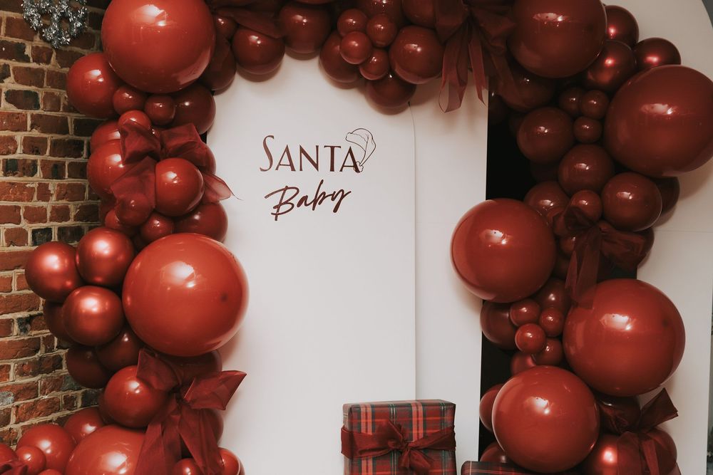Red balloon arch decorations create a festive entrance display against a white brick wall with Santa signage.