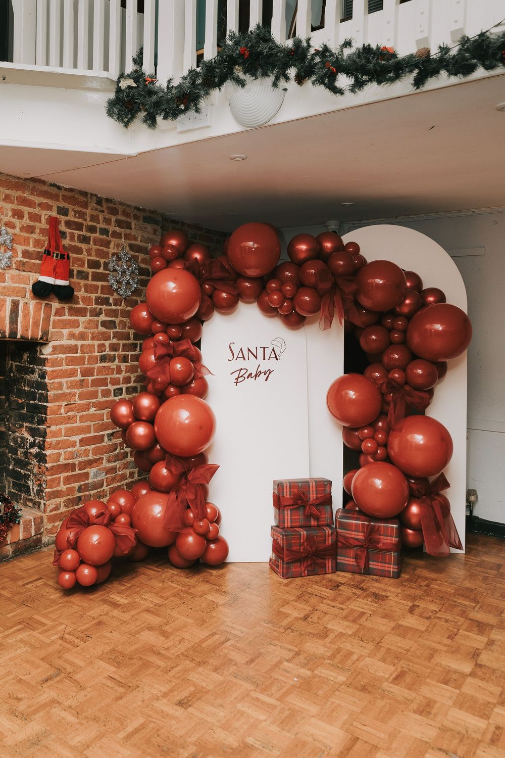 A festive Christmas setup with red balloon garland and white door marked 'Santa Baby' against exposed brick wall.