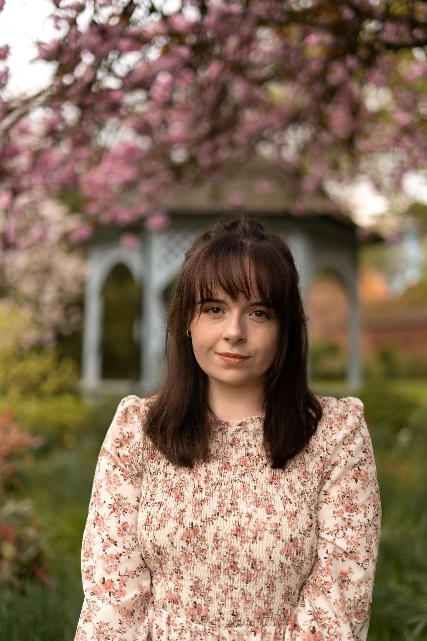 A person in a floral dress poses under blooming cherry blossom trees on a spring day.
