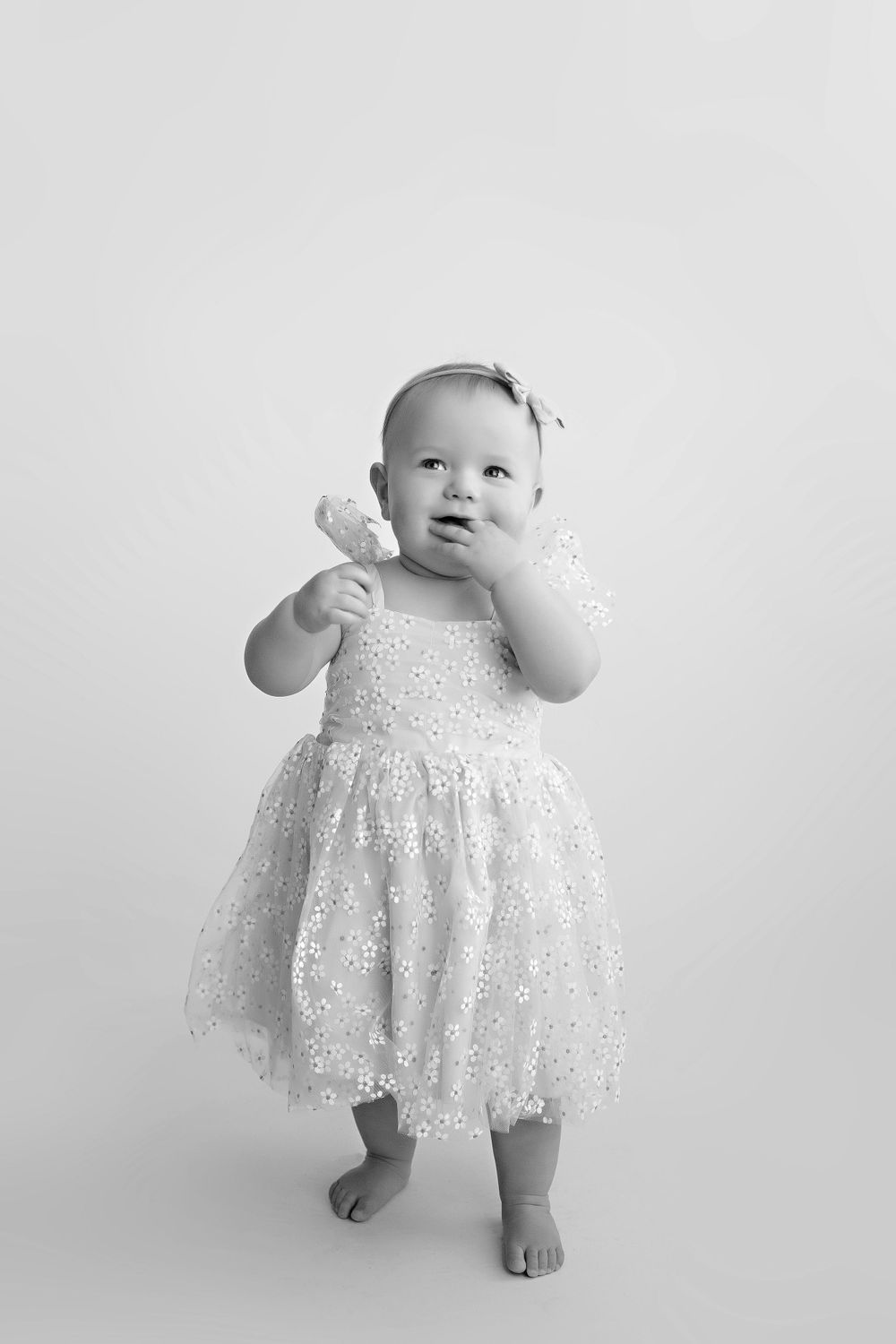 Sweet black and white portrait of infant in white lacy dress standing barefoot against light background.