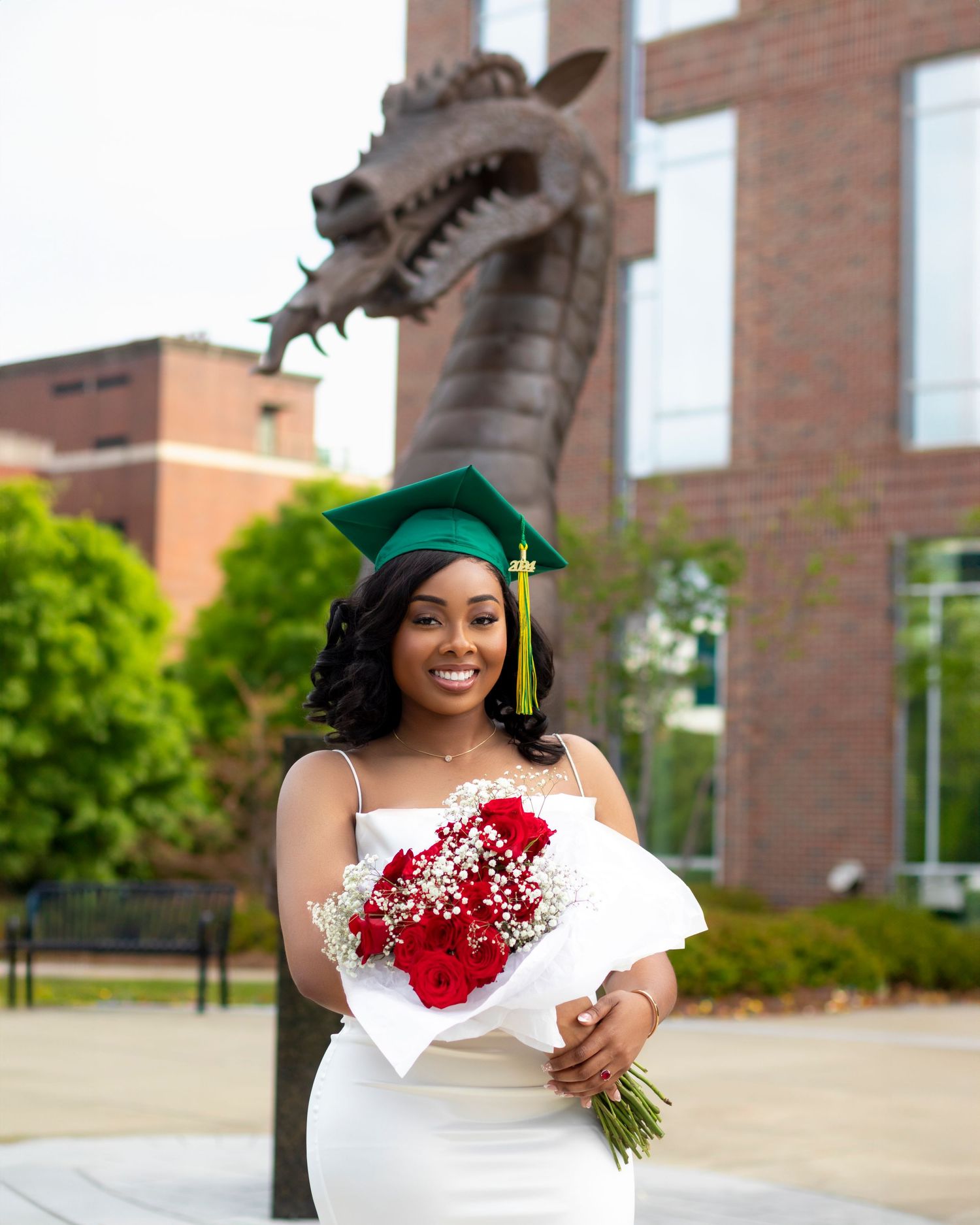 Graduate in a green cap and gown holds red roses while standing near a dragon sculpture outside a brick building.