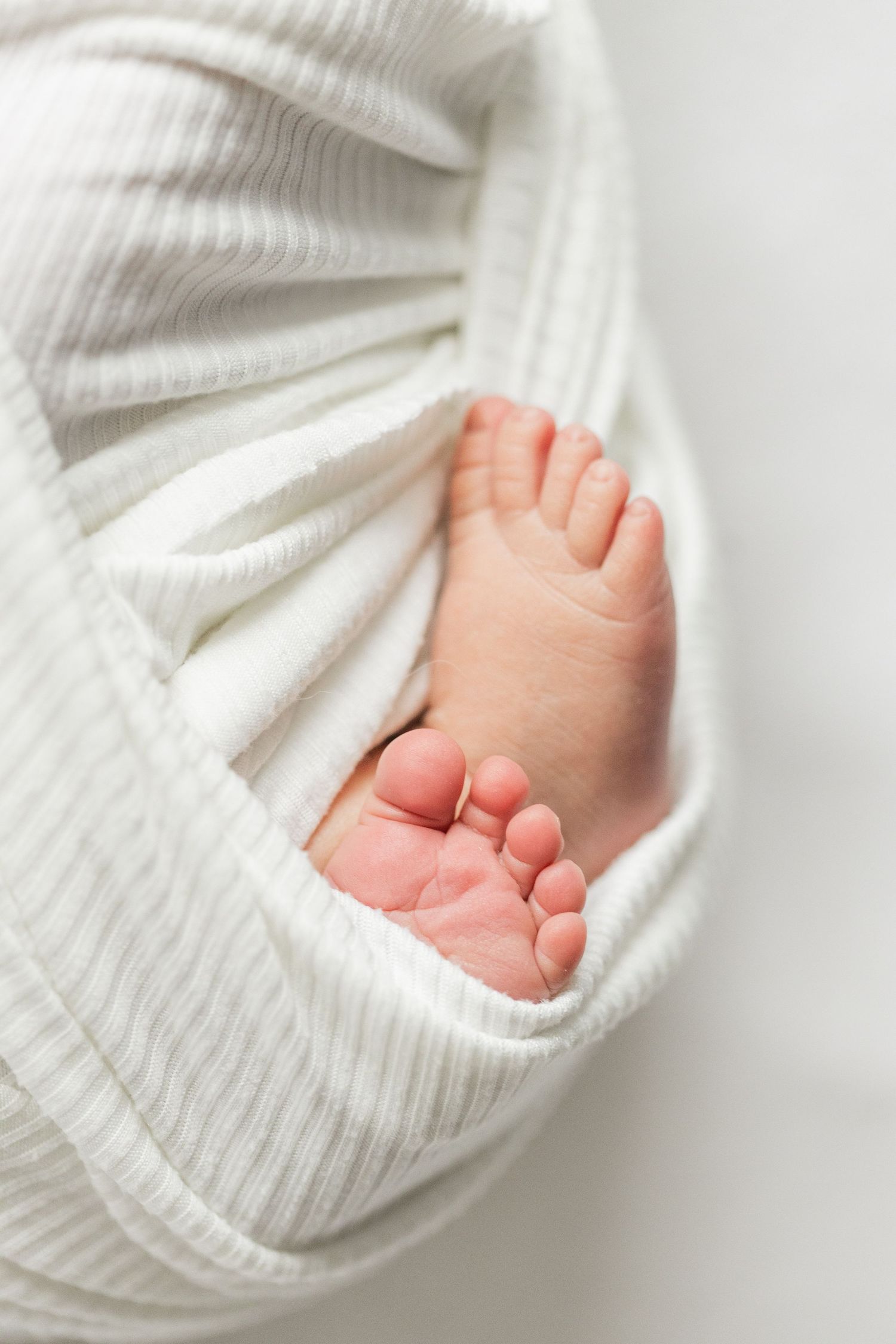 Tiny baby feet peek out from soft white knitted blanket against light background.