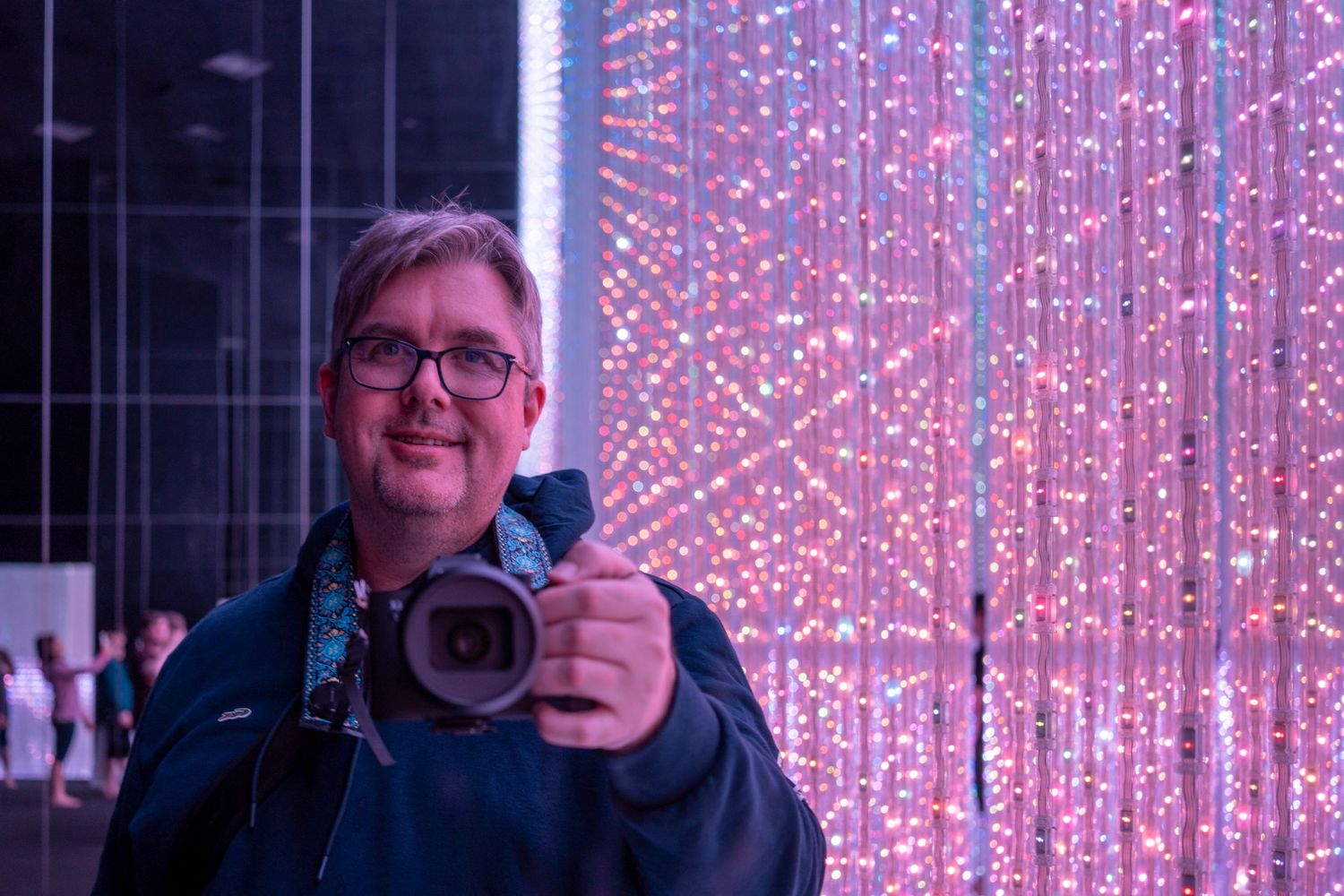 Photographer with camera poses against a backdrop of pink LED light curtains.
