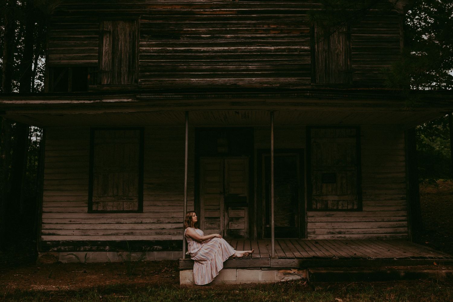 A spooky atmospheric scene of someone in a white dress sitting on the steps of an abandoned wooden cabin in the dark woods. Whitney Popp