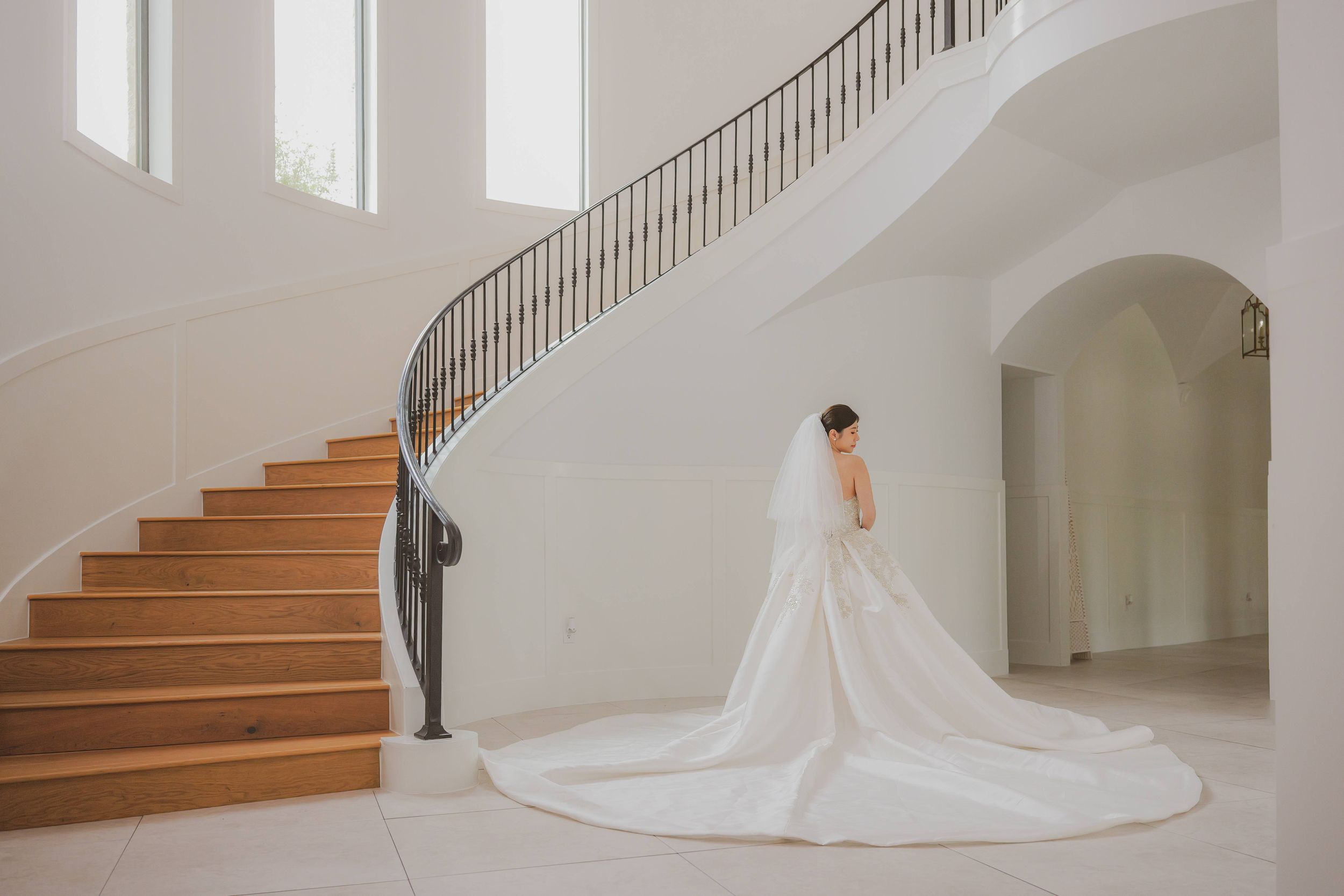 An elegant white wedding dress with a long train photographed from behind on wooden stairs in a bright interior.