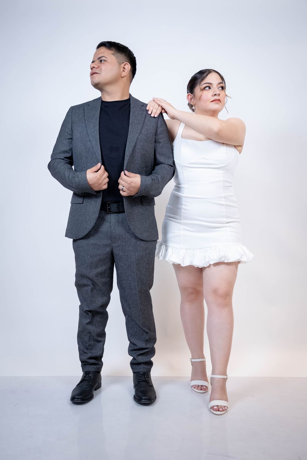 A couple in formal attire poses playfully with one in gray suit and other in white dress against light background.
