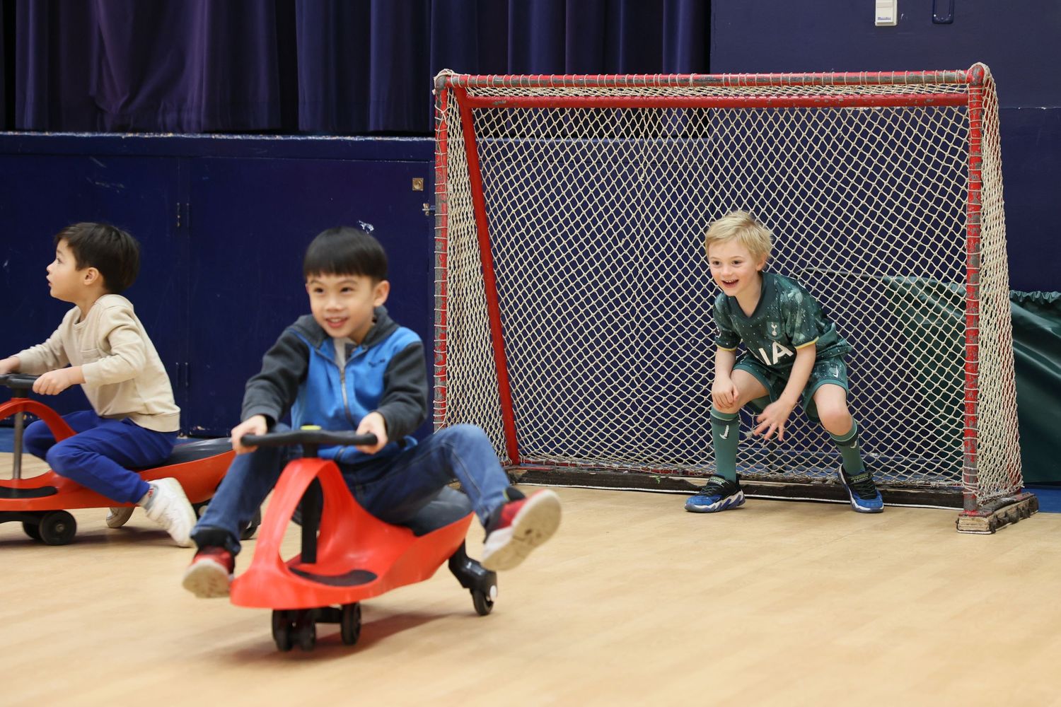 Children play with ride-on toys in a gym while one child sits in a sports goal net. Event and Birthday Photography by Smilein.
