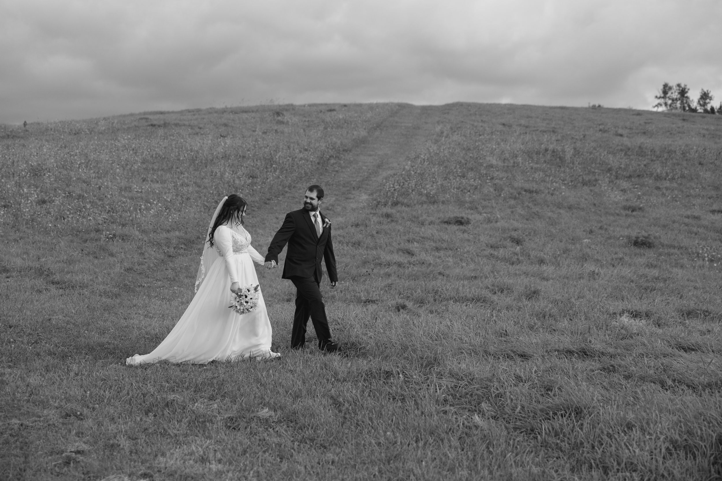 A bride and groom walk hand in hand across a windswept hilltop under moody skies in black and white.