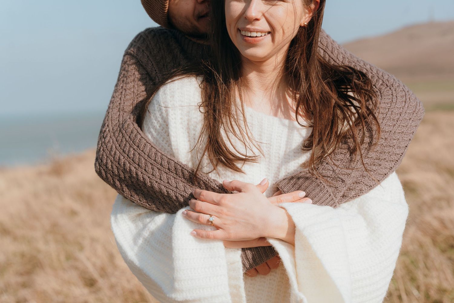 A couple embraces warmly on a grassy hillside near the ocean, wearing cozy knit sweaters.