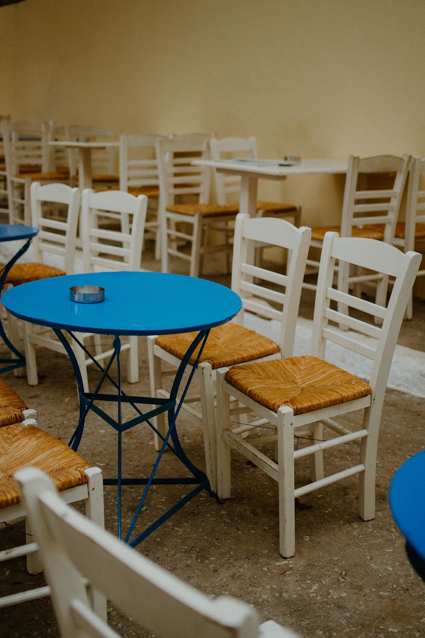 White wooden chairs with woven seats surround bright blue bistro tables in an empty cafe setting.