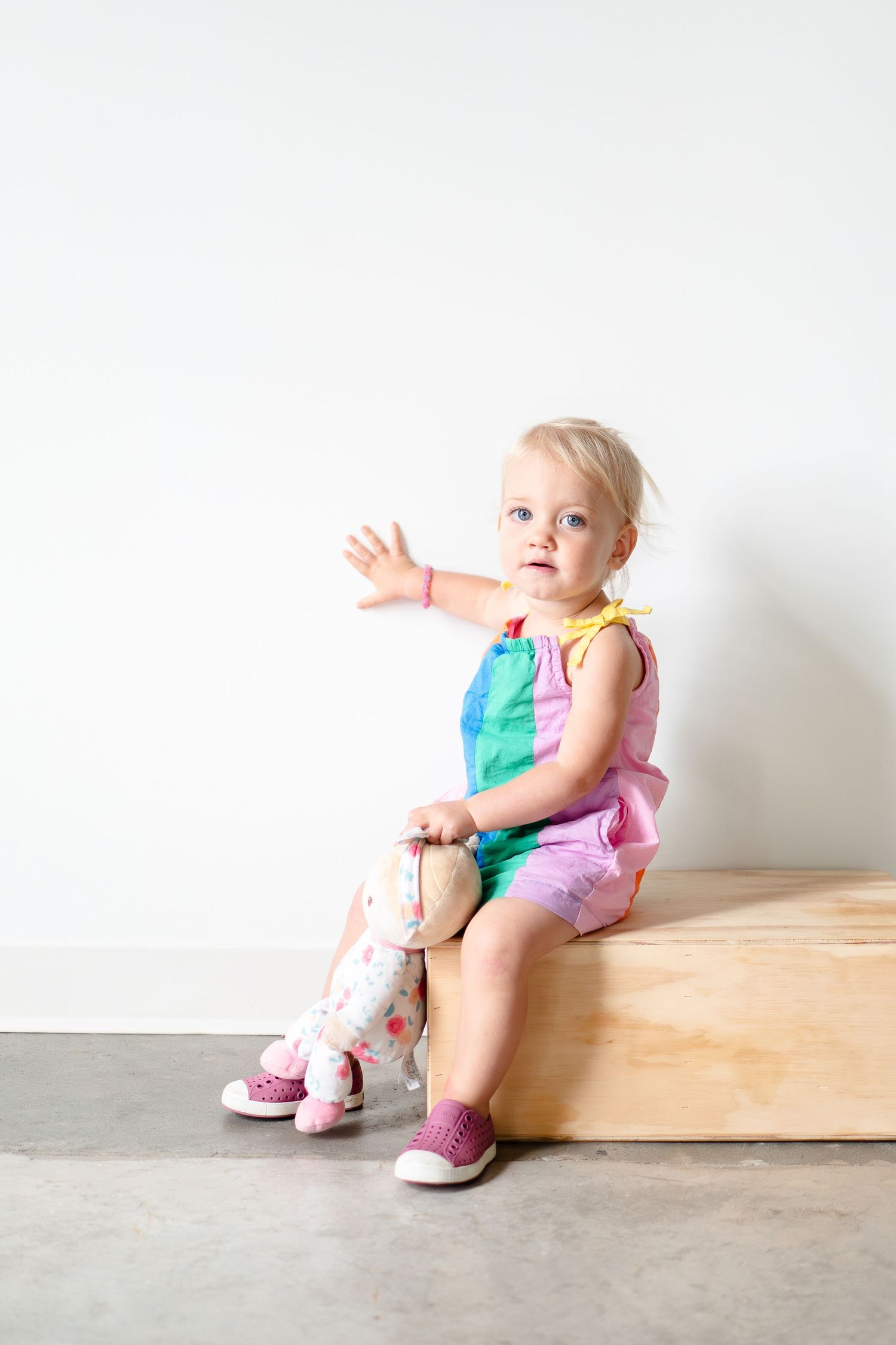 Small child in a colorful outfit sits on wooden platform holding stuffed animal toy against white wall.