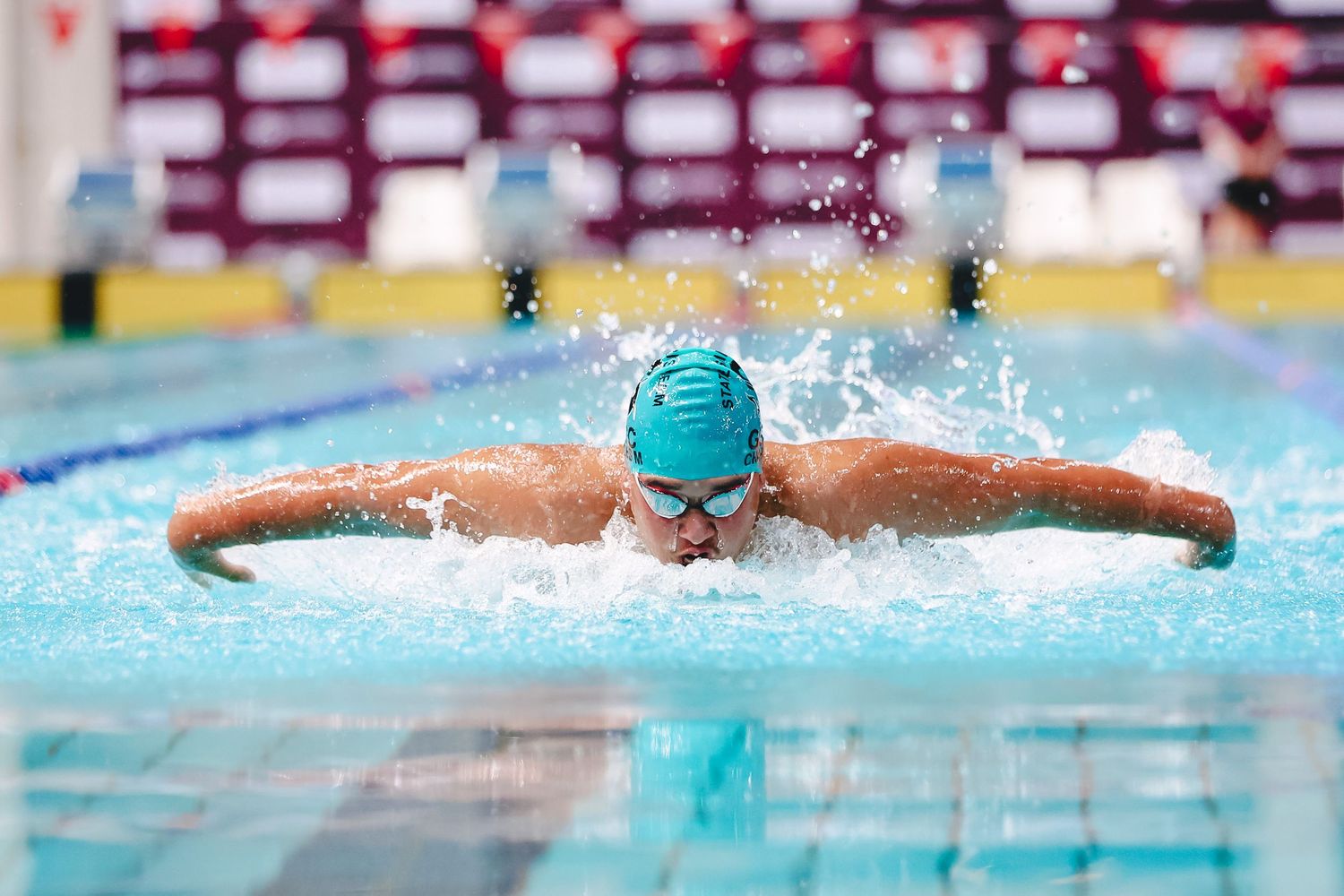 A swimmer performs the butterfly stroke in a competition pool, creating splashes in the bright blue water.