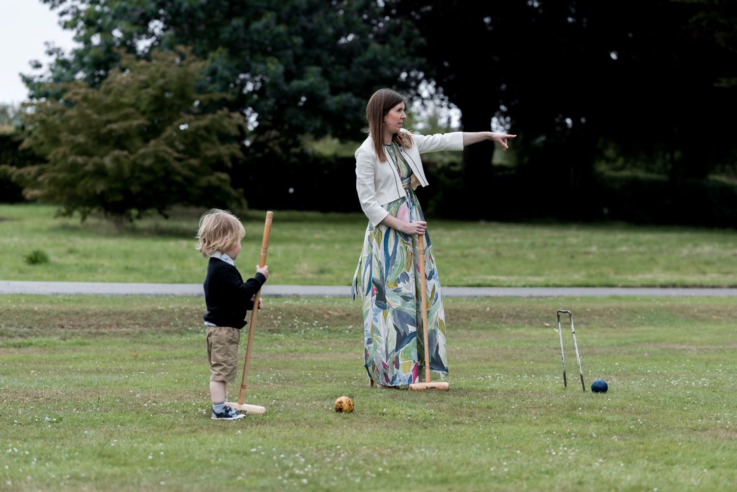 Two people playing croquet on a green lawn with trees in the background on a sunny day.