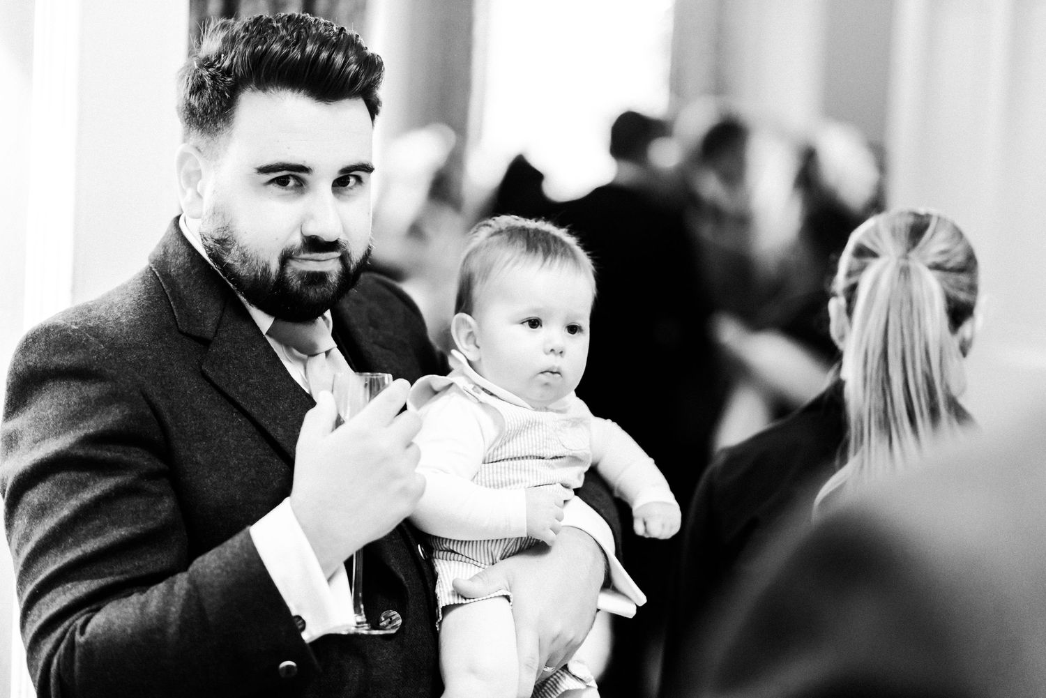 A well-dressed man in a suit holding a baby in formal attire at an event, captured in black and white.