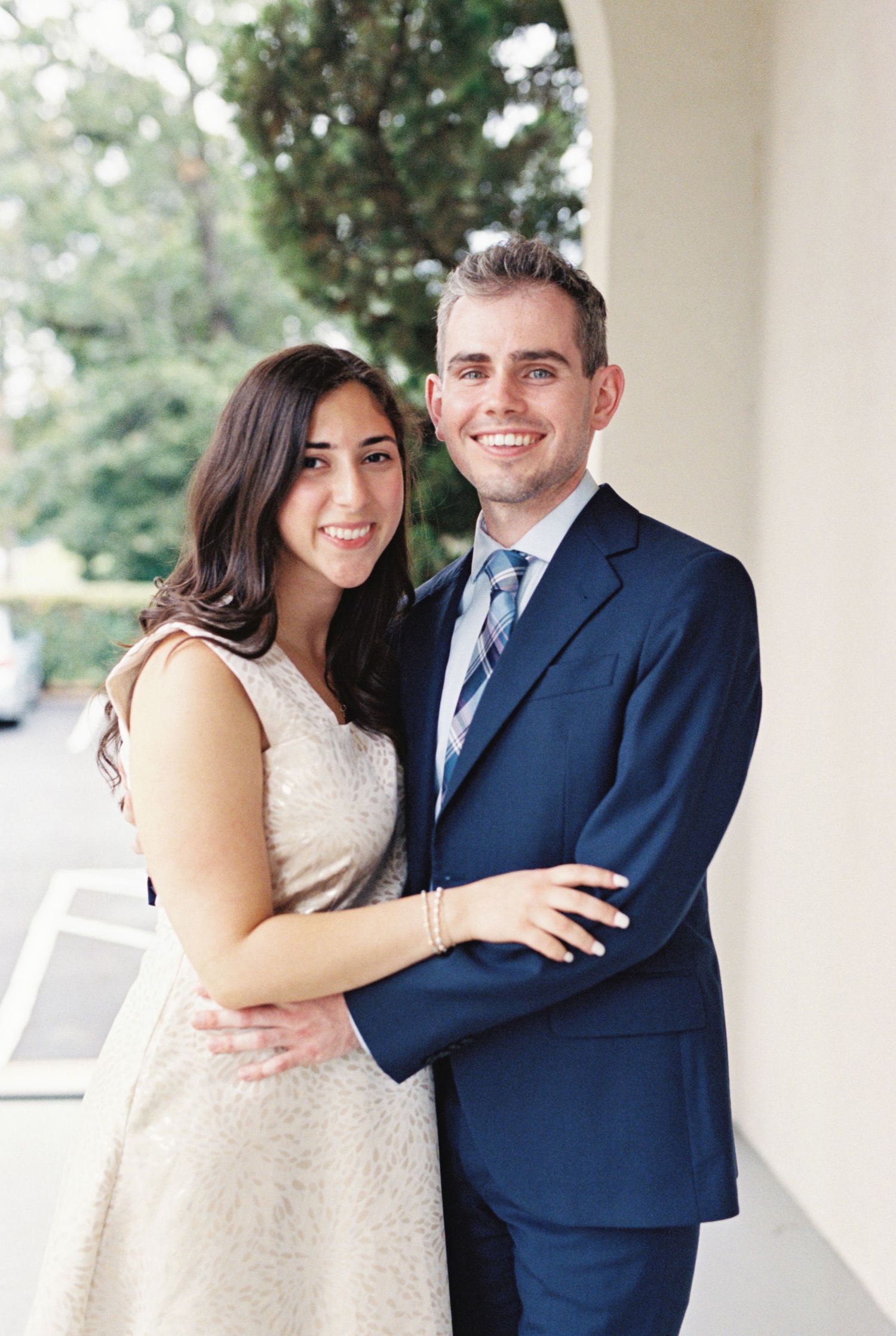 A couple in formal attire embracing on a covered porch, with the woman in white and man in navy suit.